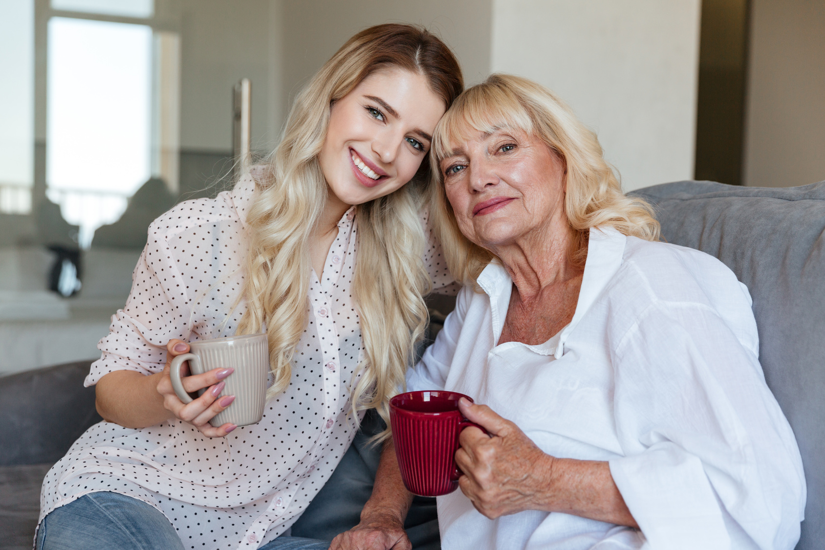 Picture of smiling young lady sitting at home with her grandmother drinking tea. Looking camera.